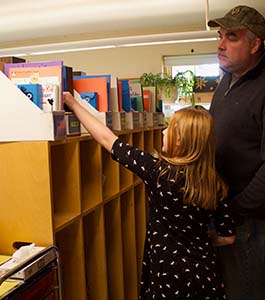Girl reaching for book on shelf with father